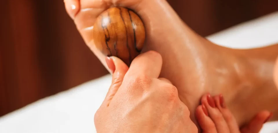 Técnica de reflexología podal con una bola de madera, donde se masajea el pie para aliviar el estrés y mejorar la circulación.