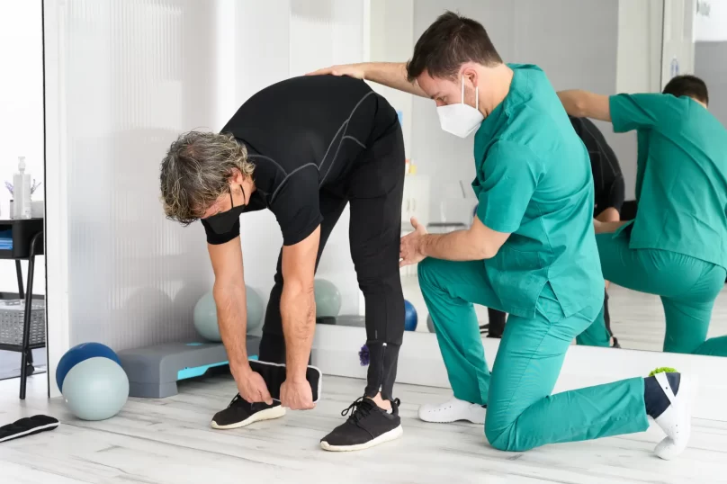 Fisioterapeuta asistiendo a un paciente mayor en una sesión de rehabilitación, enfocándose en estiramientos y ejercicios de movilidad en un entorno clínico.