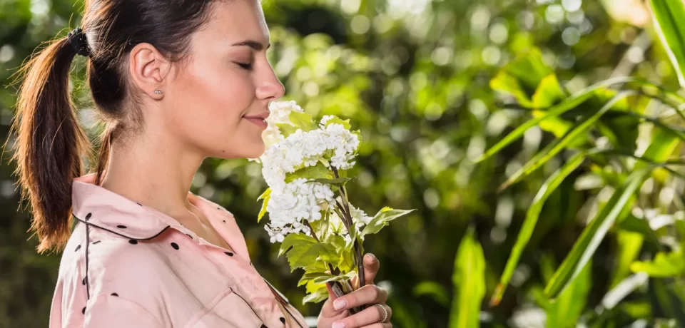 Mujer oliendo una flor blanca en un entorno natural, rodeada de vegetación. La imagen transmite tranquilidad y conexión con la naturaleza.
