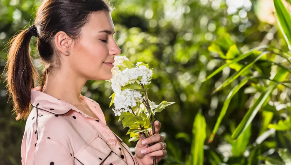 Mujer oliendo una flor blanca en un entorno natural, rodeada de vegetación. La imagen transmite tranquilidad y conexión con la naturaleza.