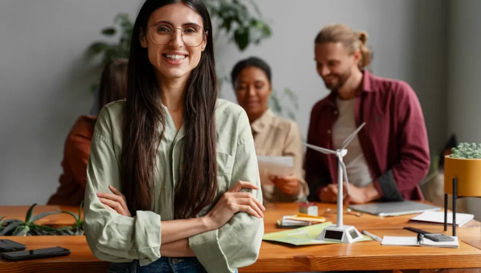 Mujer sonriente con camisa verde en una reunión de trabajo, rodeada de colegas, con un modelo de turbina eólica en la mesa.